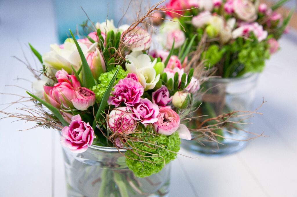Zwei Glasvasen mit frischen, farbenfrohen Blumensträußen stehen auf einem hellen Tisch. Die Sträuße bestehen aus rosa, weißen und grünen Blumen sowie verschiedenen grünen Blättern und Zweigen.